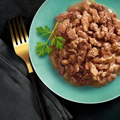 Bord met stukjes natvoer in saus, gegarneerd met een takje peterselie. Gouden vork en zwarte servet zichtbaar naast het bord.