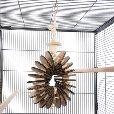 Jouet suspendu en corde, bois et carton ondulé, accroché dans une cage à oiseaux avec perchoirs en bois visibles.