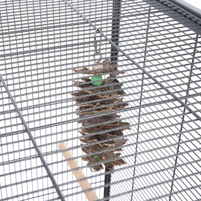 Jouet suspendu en écorce et perles vertes accroché à la grille d'une cage pour oiseaux, avec un crochet métallique visible.