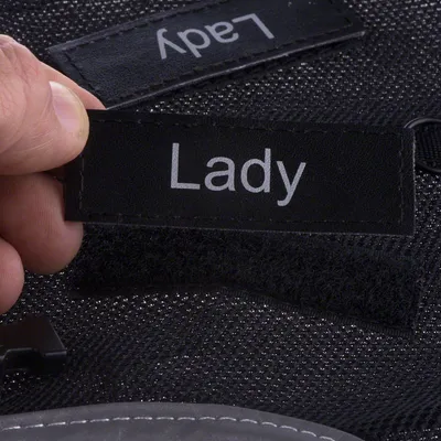 Sort velcromærke med teksten 'Lady', holdes af en hånd. Baggrund af sort stof med flere mærker synlige.