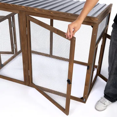 Enclos en bois avec toit en métal ondulé, porte grillagée ouverte tenue par une main, structure à mailles métalliques visibles.