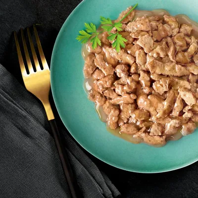 Chunks of wet pet food in gravy served on a green plate with a parsley garnish, shown next to a gold fork on a dark cloth napkin.