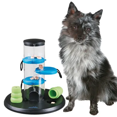 Куче до интерактивна играчка за лакомства с три прозрачни цилиндъра, сини платформи и зелени гумени елементи върху черна основа. Видими са парченца лакомства.