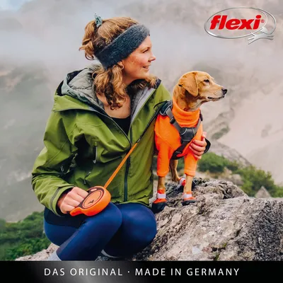 Flexi. Het origineel - Made in Germany. Vrouw houdt een Flexi-rollijn, hond draagt oranje tuig.