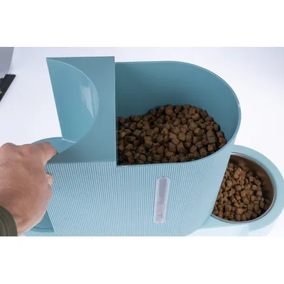 Hand bedient einen blauen Futterspender, gefüllt mit Trockenfutter. Neben dem Spender befindet sich eine Schüssel mit Trockenfutter.