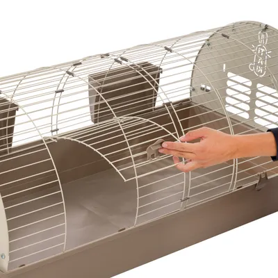 Main ouvrant la porte d'une cage pour petits animaux en métal et plastique beige, deux mangeoires visibles à l'intérieur.