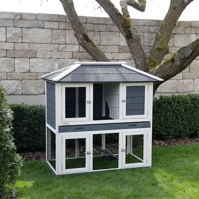 Jaula de dos pisos para pequeños animales, blanca y gris, con puertas de rejilla, apta para exterior. Techo con cubierta negra, rodeada de jardín.