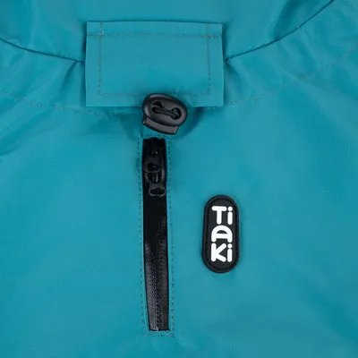 Détail d’un vêtement ou accessoire en tissu bleu avec fermeture éclair noire, cordon de serrage et écusson visible portant la marque Tiaki.
