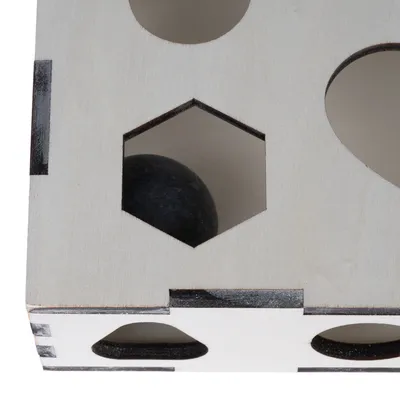 Coin d’un cube en bois clair avec ouvertures de formes variées, dont un hexagone laissant voir une balle noire à l’intérieur. Matériau visible : bois.