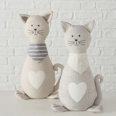 Zwei dekorative Stoffkatzen mit Herzmotiv auf dem Bauch, eine mit gestreiftem Halstuch und die andere mit floralem Halstuch. Beide in neutralen Farben vor weisser Ziegelwand.