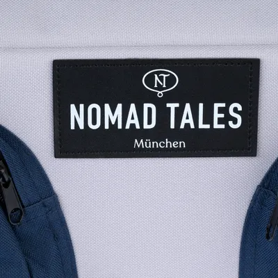 Nomad Tales München. Metka marki na szarej tkaninie z niebieskimi akcentami.