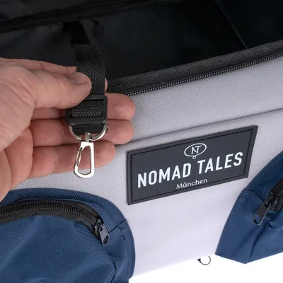 Nomad Tales München torba z szarego materiału, niebieskimi kieszeniami i czarnym karabińczykiem. Ręka trzyma karabińczyk do mocowania.