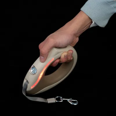 Mano che tiene un guinzaglio per cani beige con illuminazione LED e moschettone in metallo.