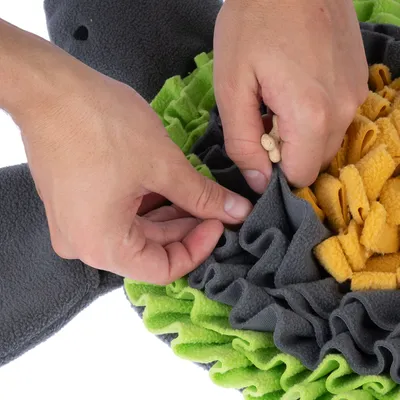 Hände verstecken Leckerlis in einem Schnüffelteppich aus grauem, grünem und gelbem Stoff. Demonstration der Nutzung des interaktiven Spielzeugs.