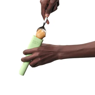 Hand hält grünes Gummi-Spielzeug mit Textur, während ein Löffel Erdnussbutter darauf verteilt wird.