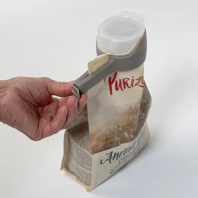 Roka prikazuje, kako je siv pokrov z dozirno odprtino nameščen na embalažo Purizon Ancient Grains Chicken.