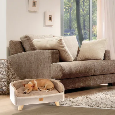 Hund schläft in einem beigen Hundebett mit Holzfüssen vor einem braunen Sofa in einem Wohnzimmer.