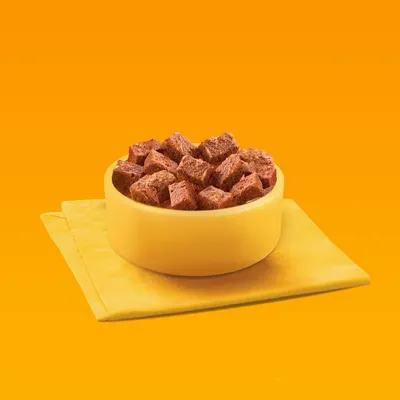 Gelbe Schale mit braunen Fleischwürfeln auf gelber Serviette vor orangefarbenem Hintergrund.