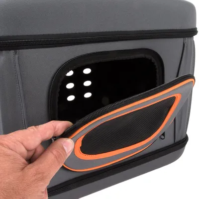 Vue rapprochée d’une caisse de transport grise en tissu avec ouverture zippée, filet noir et bordure orange, main ouvrant la porte latérale ajourée.