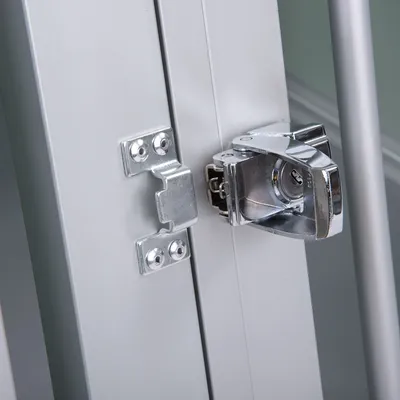 Metalowy zamek i zaczep na drzwiach lub klatce, widoczne elementy montażowe i konstrukcja z aluminium.