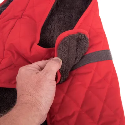 Detailní záběr na červenou prošívanou psí bundu s měkkou podšívkou. Ruka ukazuje otvor a pásek na suchý zip.