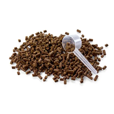 Brązowe granulki karmy sypane na białym tle, przezroczysta plastikowa miarka częściowo wypełniona granulkami widoczna po prawej stronie.