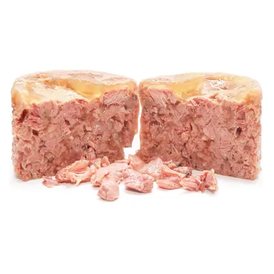 Cibo umido per animali tagliato a metà, consistenza compatta con pezzi visibili di carne rosa e gelatina superficiale, alcuni bocconcini sparsi davanti.