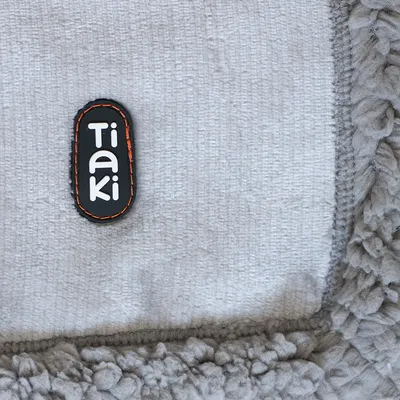 Étiquette avec le nom de la marque Tiaki sur tissu gris à bordure en fausse fourrure bouclée.