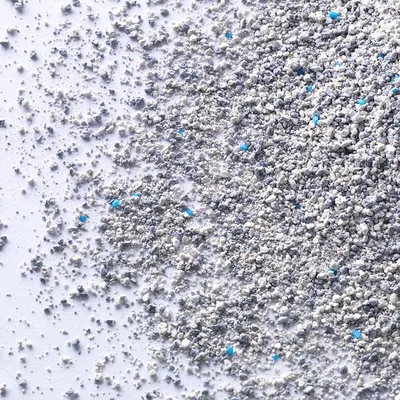 Arena aglomerante para gatos de color gris claro con pequeños gránulos azules visibles, extendida sobre una superficie blanca.