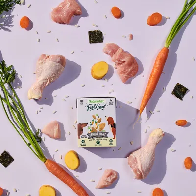Naturediet Feel Good Senior Light food pack surrounded by visible ingredients including raw chicken, carrots, potatoes, rice grains and seaweed pieces.