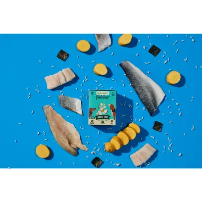 Product image showing a box of Naturediet Feel Good White Fish surrounded by visible ingredients including fish fillets, potato slices, rice grains and seaweed pieces on a blue background.