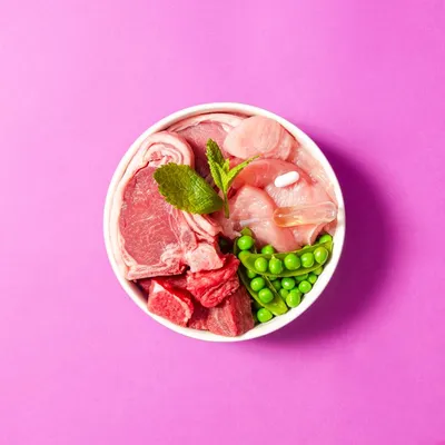 Bowl containing raw meat cuts, green peas, a mint leaf, a white tablet and a clear liquid capsule, showing key ingredients of pet food.