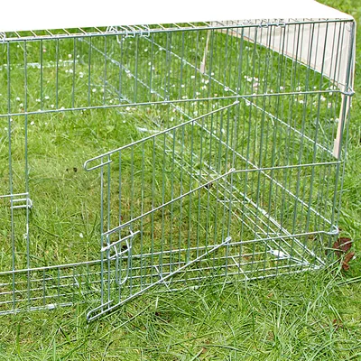 Enclos rectangulaire en métal avec porte ouverte, posé sur de l’herbe. Structure grillagée, toit partiellement couvert par une toile claire.