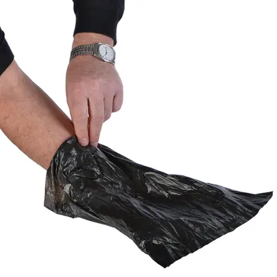 Main d’adulte insérant la main dans un sac en plastique noir, manche relevée, montre visible au poignet gauche. Sac sans marque ni texte apparent.