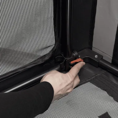 Hånd peger på rød lås til fastgørelse af metalramme med netstof. Detaljevisning af montering af dyretransportbur.