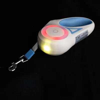 Laisse rétractable blanche et bleue avec poignée, bouton d’alimentation visible, anneau lumineux rouge, lumière LED jaune allumée, mousqueton métallique pour attacher au collier.