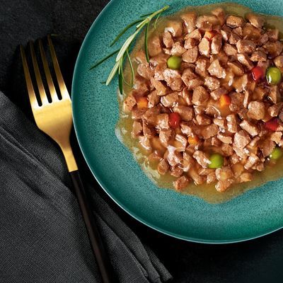 Portie natvoer met stukjes vlees, erwten en wortel in saus op een groen bord, gegarneerd met rozemarijn. Gouden vork en zwarte servet zichtbaar naast het bord.