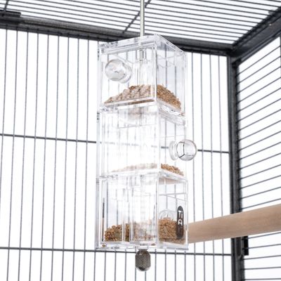 Przezroczysty podajnik na karmę z tworzywa sztucznego z kilkoma komorami, wypełniony ziarnem. Zamontowany wisząco w klatce dla ptaków.