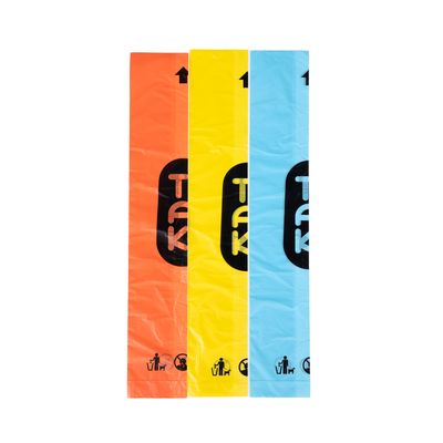 Tre rotoli di sacchetti per deiezioni canine in arancione, giallo e blu con logo TAK. Simboli di smaltimento e cane visibili.
