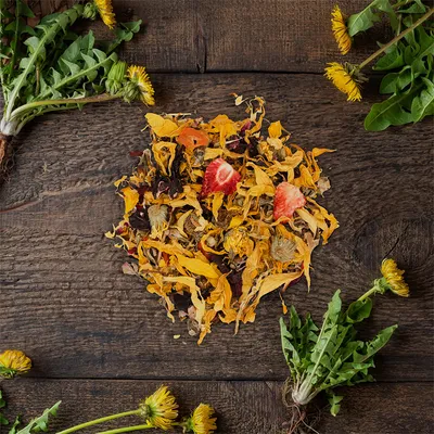 Mélange de fleurs séchées jaunes et rouges avec morceaux de fraise, entouré de pissenlits frais sur une table en bois foncé. Aucun emballage visible.