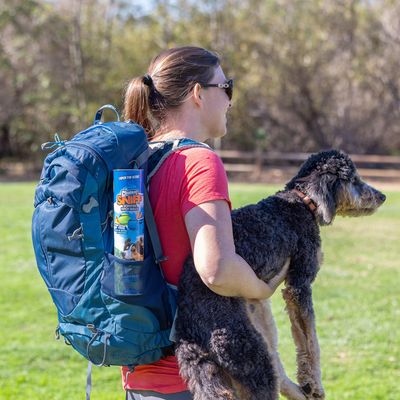 Zaino con il prodotto 'Catch the Scent Crunchy Sniff' nella tasca laterale, indossato da una persona che tiene un cane. Sfondo all'aperto.