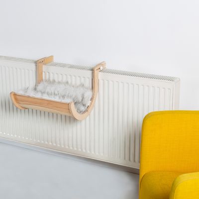 Dřevěné závěsné lehátko s šedým potahem z umělé kožešiny, připevněné k radiátoru. V popředí žluté křeslo.