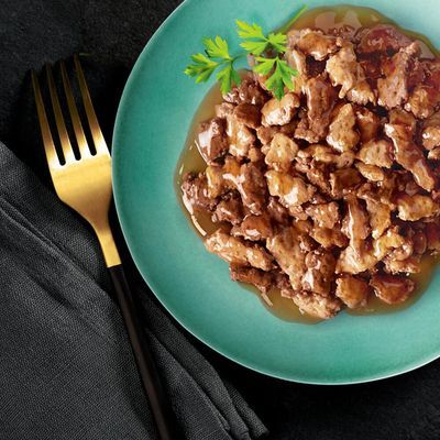 Bord met stukjes natvoer in saus, gegarneerd met peterselie, op een groene schaal naast een gouden vork en zwarte servet. Merknaam of productinformatie niet zichtbaar.