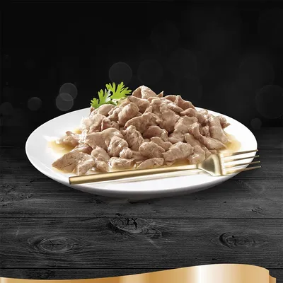Chunks of meat in gravy on a white plate with a gold fork and parsley garnish, shown on a dark wooden surface. No visible brand or product name.