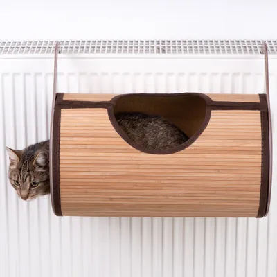 Cama para gatos de madera con borde de tela marrón, fijada a un radiador. Gato asomando por la abertura.