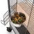 Gamelle en métal accrochée à une cage, remplie de graines, maïs et croquettes vertes pour oiseaux ou petits animaux. Support métallique visible sur la porte de la cage.