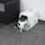 Weisse Katzentoilette mit grüner Schublade und Klappe, Katze verlässt die Toilette. Gezeigt auf grauem Fliesenboden neben einem schwarzen Rattanstuhl.