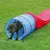 Perro grande saliendo de un túnel de agilidad azul y rojo para perros, colocado sobre césped al aire libre. El túnel es de tela con refuerzos visibles.