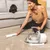 Woman using a pet hair vacuum cleaner on a carpet while a black dog sits on the sofa.