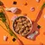 Bowl of cubed pet food surrounded by raw chicken pieces, carrots, rice grains and seaweed on an orange background, showing visible ingredients used in the product.